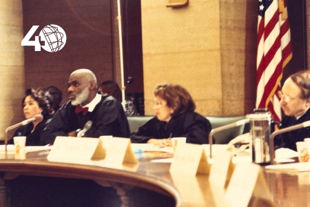 [ai] A panel of four judges seated at a conference table, with various name tags visible. They are wearing judicial robes and there is an American flag in the background, suggesting a formal legal setting.